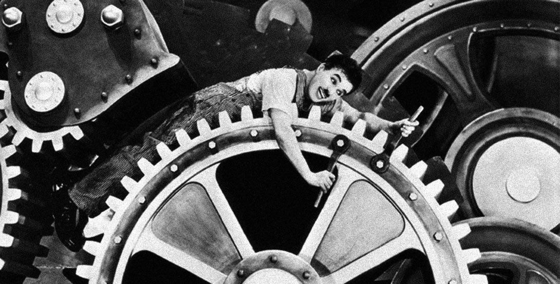 Imagen de Charles Chaplin en la película Tiempos Modernos. Está enredado entre ruedas dentadas de un mecanismo.